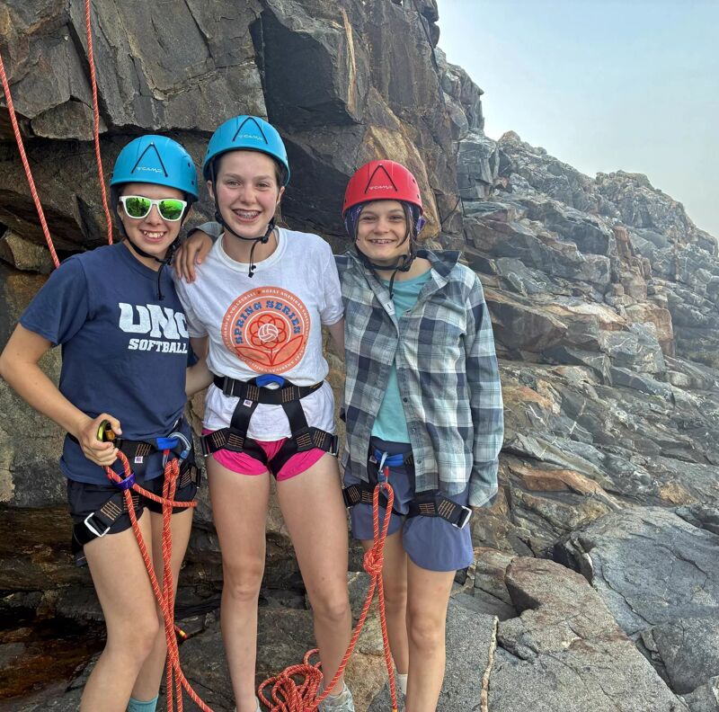 Three young women are standing close together, likely on a rocky terrain, possibly getting ready for or just finishing rock climbing. They are all wearing helmets and harnesses, indicating their involvement in climbing activities. The woman on the left is wearing sunglasses and a blue t-shirt, the one in the center has a white t-shirt, and the woman on the right is wearing a red helmet and a plaid shirt. Ropes are visible, further suggesting they are equipped for climbing.